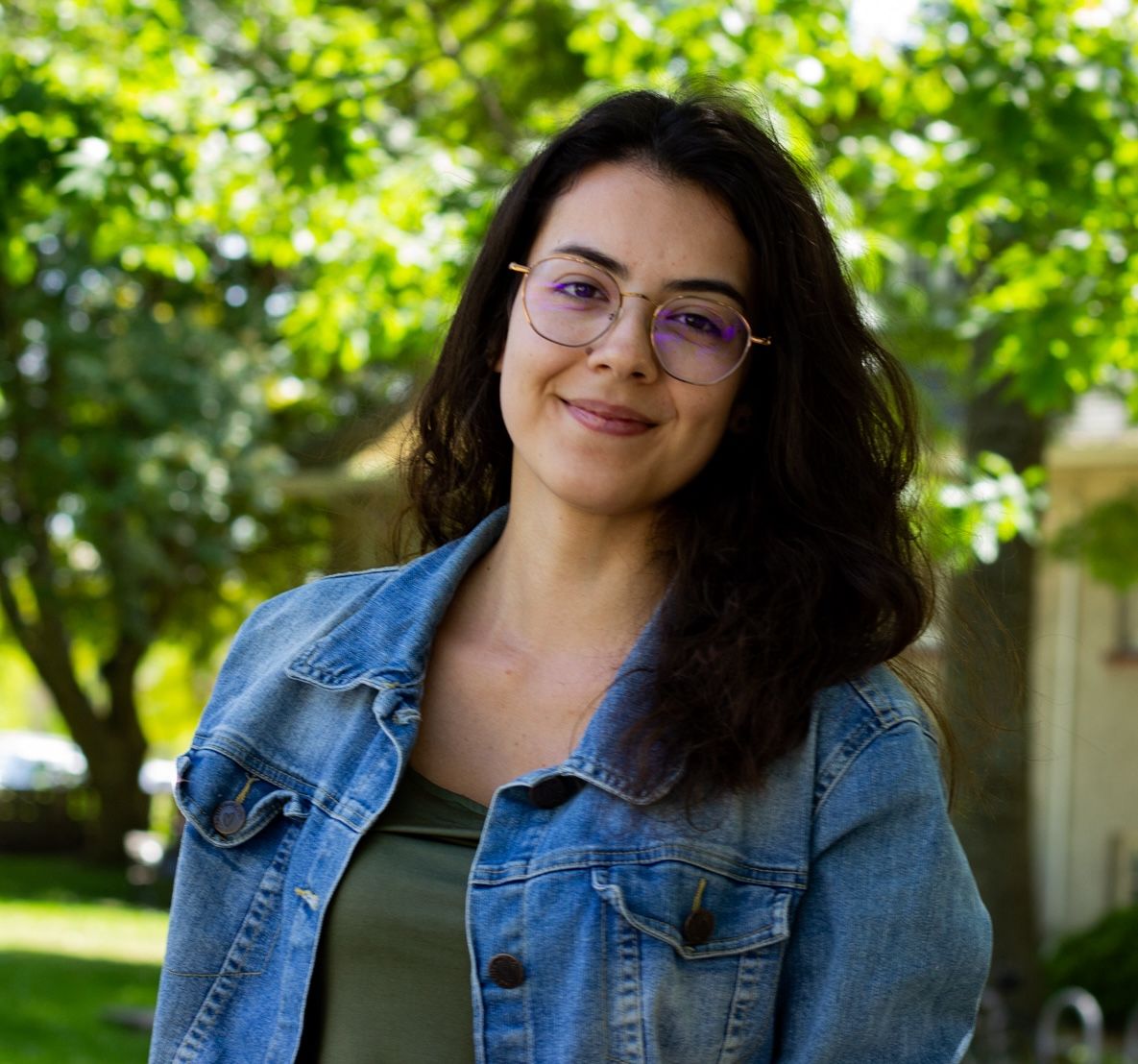 Young woman wearing glasses, a demin jacket and green shirt smiling with mouth closes. Nature background.